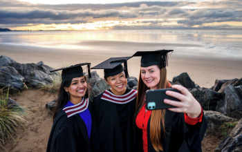 Three females dressed in graduation robes pose for a selfie with a beach in the background