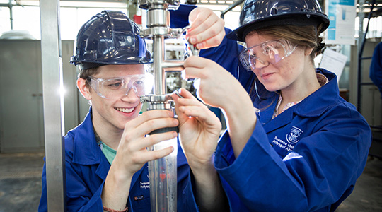 One male and one female student operating a mechanical instrument. They are wearing blue overalls, blue hard hats and protective glasses.