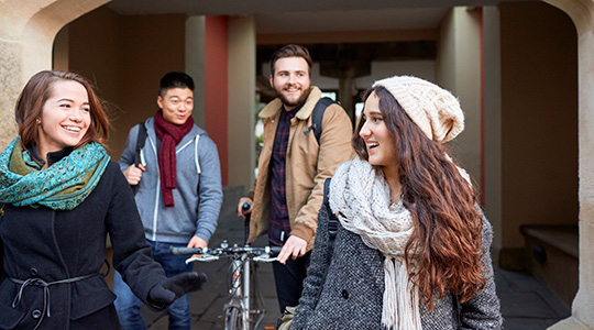 Four students walking through the grounds of Singleton Park Campus. There are two females who are smiling at each other. There are two males in the background, one of them is pushing a bike.
