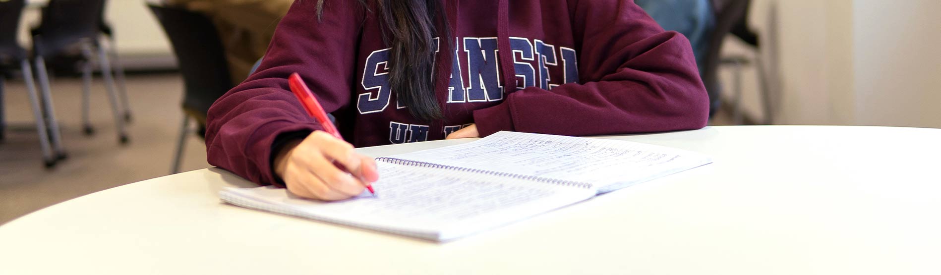A person in a red Swansea University hoody writing revision notes.
