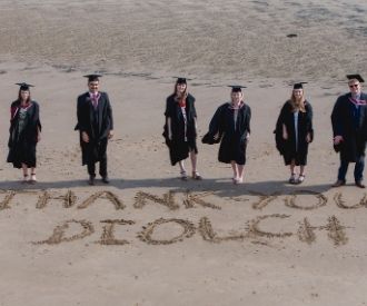 A group of Swansea graduates in caps and gowns. They're standing on the beach, with 'Thank you' written in the sand.