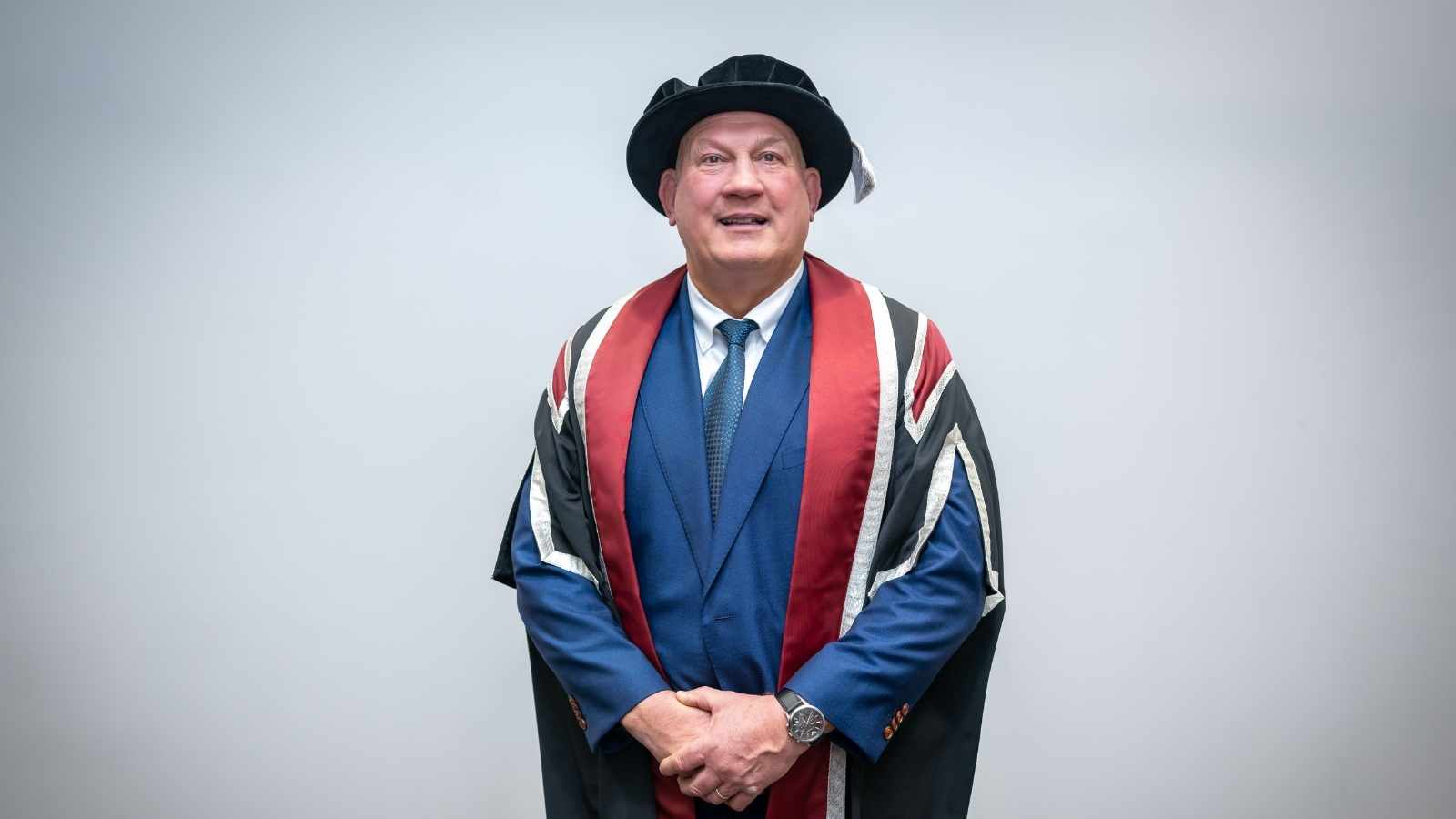 Man wearing cap & gown standing in front of a blank wall smiling at the camera