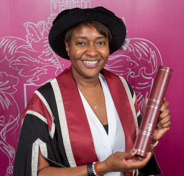 Lady wearing a cap & gown holding a scroll & smiling from the camera