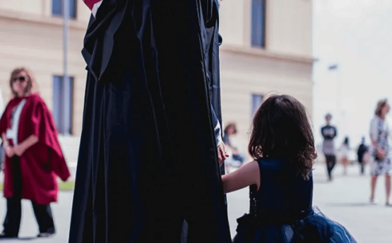 A graduate holding hands with their dark haired child.