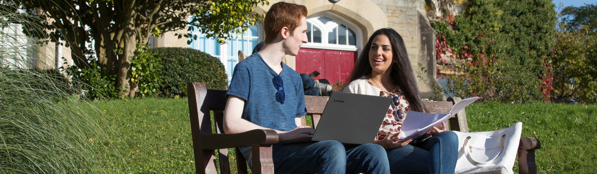 One male on a laptop and one female with a booklet, sitting on a bench outside the Abbey on Singleton Campus surrounded by trees. 