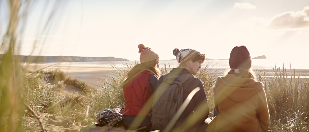 Three students in coats and hats sitting on a beach and talking