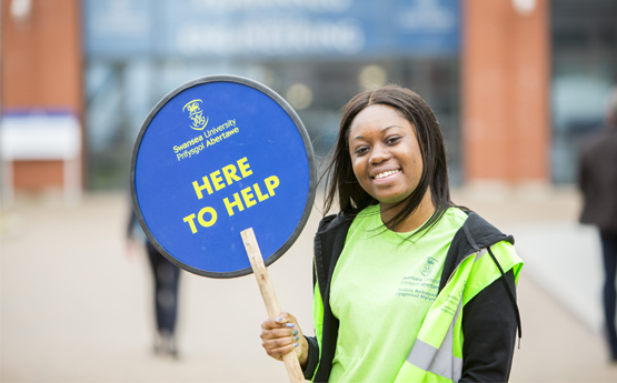 Female Student ambassador in a hi-visibility jacket with a 'here to help' sign