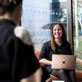 An image of a student talking to another student working at a laptop.