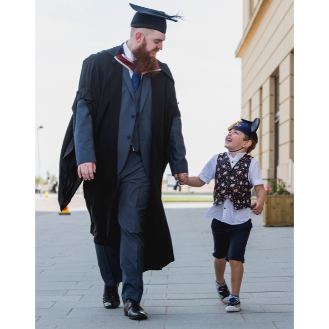 An adult student in cap and gown walks hand in hand with a child on Bay campus at Swansea University 