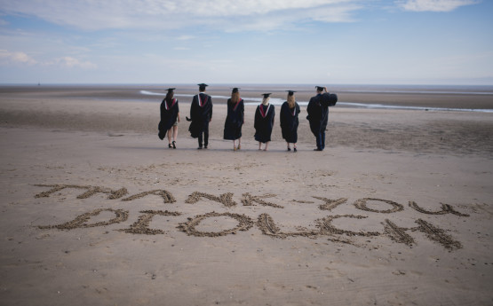 graduates in cap and gowns, walking on the beach with the words Thank you/Diolch written in the sand