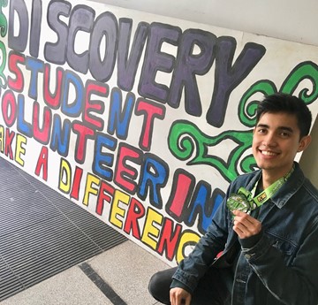 Kritsanaphong Sangraksa kneeling beside a hand painted Discovery sign and holding up a medal.