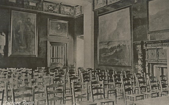 A lecture theatre in the Abbey in the 1920s. Rows of seats in a grand room with paintings and an ornate fireplace. 