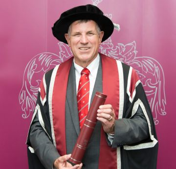 Dyn mewn cap a gwn yn dal scrol yn gwenu i'r camera