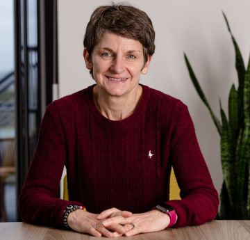 Menyw yn eistedd wrth ddesg gyda'i dwylo o'i blaen yn gwenu i'r camera