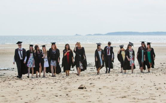 Myfyrwyr mewn gynau graddio ar y traeth