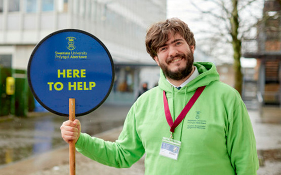 Myfyriwr mewn gwyrdd leim yn dal arwydd lolipop 'Yma i Helpu'