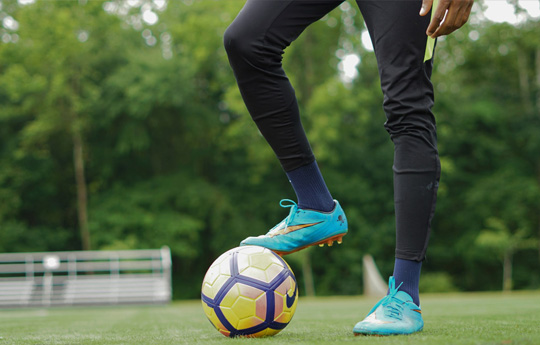 Person mewn legins du ac esgidiau pêl-droed glas gydag un droed ar bêl felen