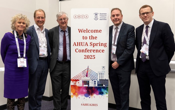 Pump o bobl wedi'u gwisgo'n drwsiadus - pedwar dyn ac un fenyw - yn sefyll ochr yn ochr â baner naid cynhadledd.