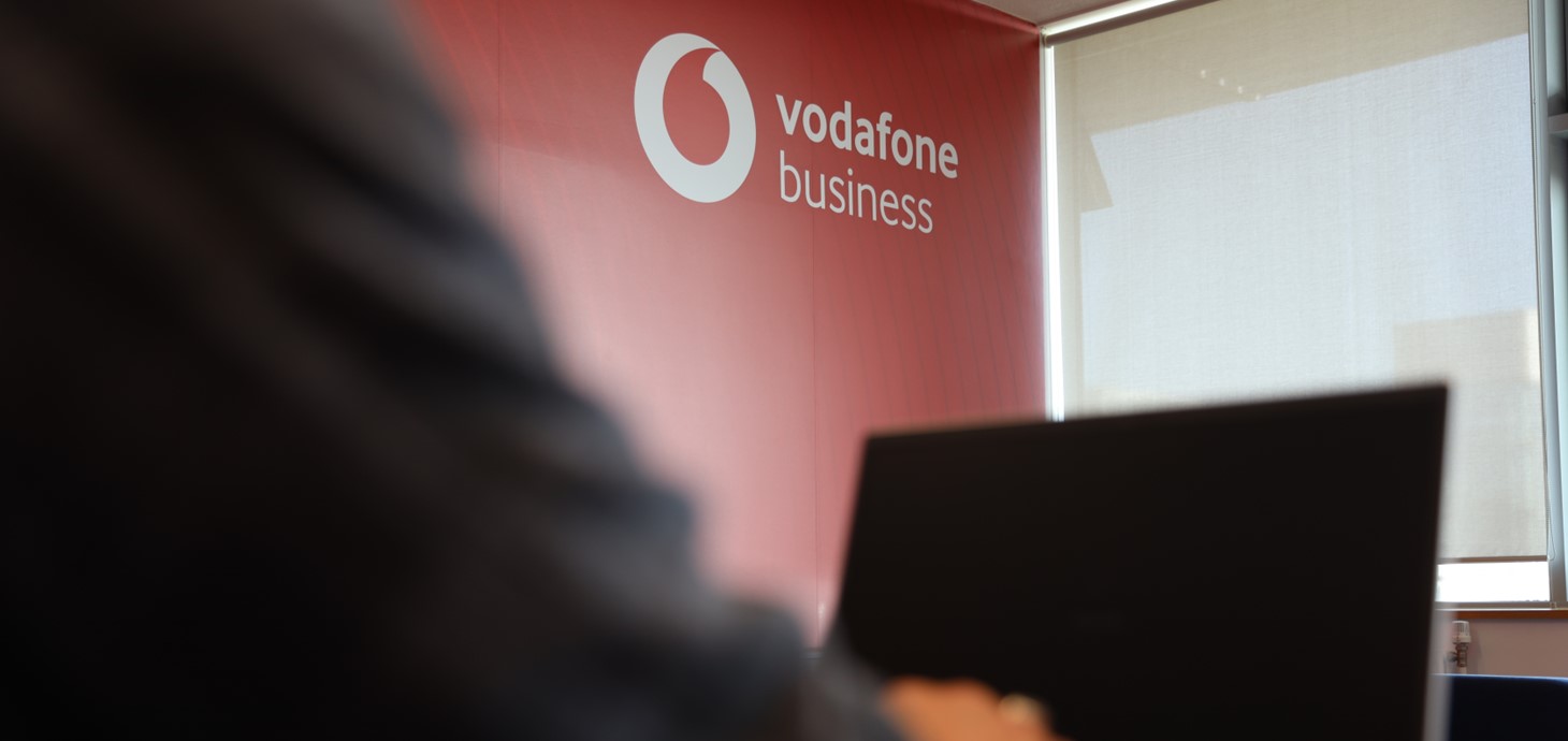 Golwg agos ar ddwylo'n teipio ar liniadur. Gwelir logo Vodafone ar y wal goch yn y cefndir. 