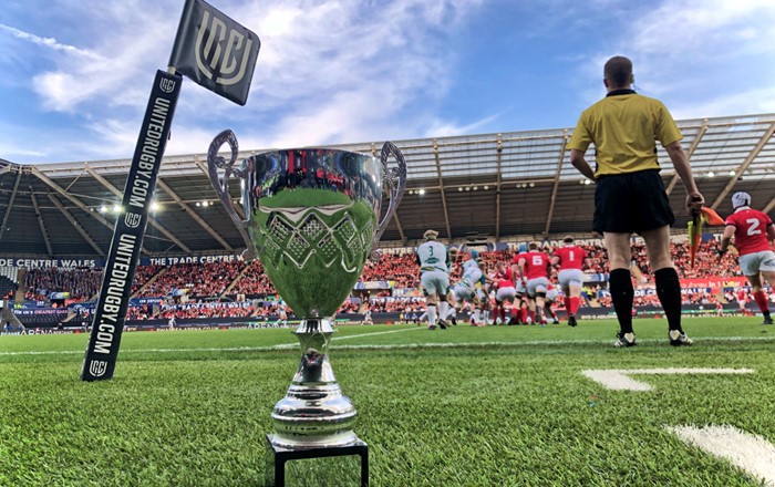 Cwpan Varsity ar lawr cae rygbi gyda chwaraewyr yn y pellter 