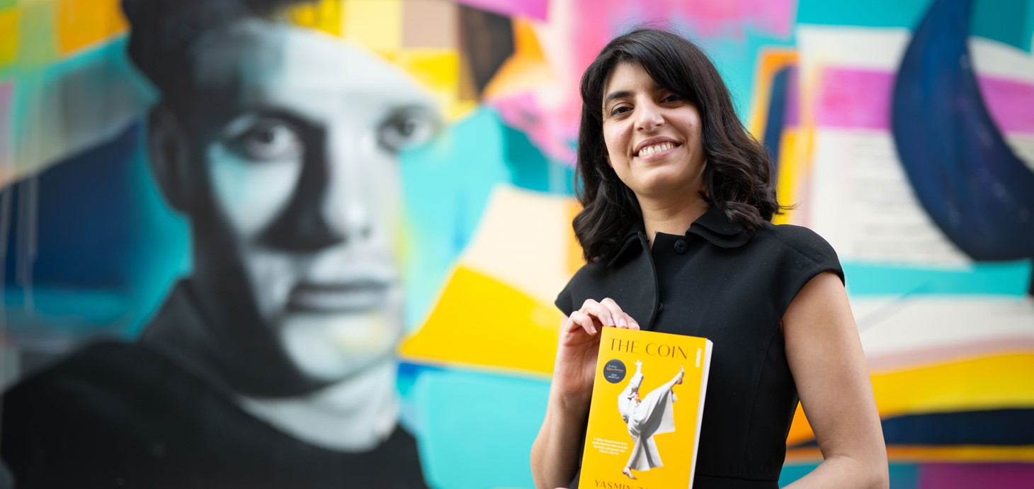 Yasmin Zaher gyda'i nofel The Coin yn sefyll o flaen murlun lliwgar o wyneb Dylan Thomas. 