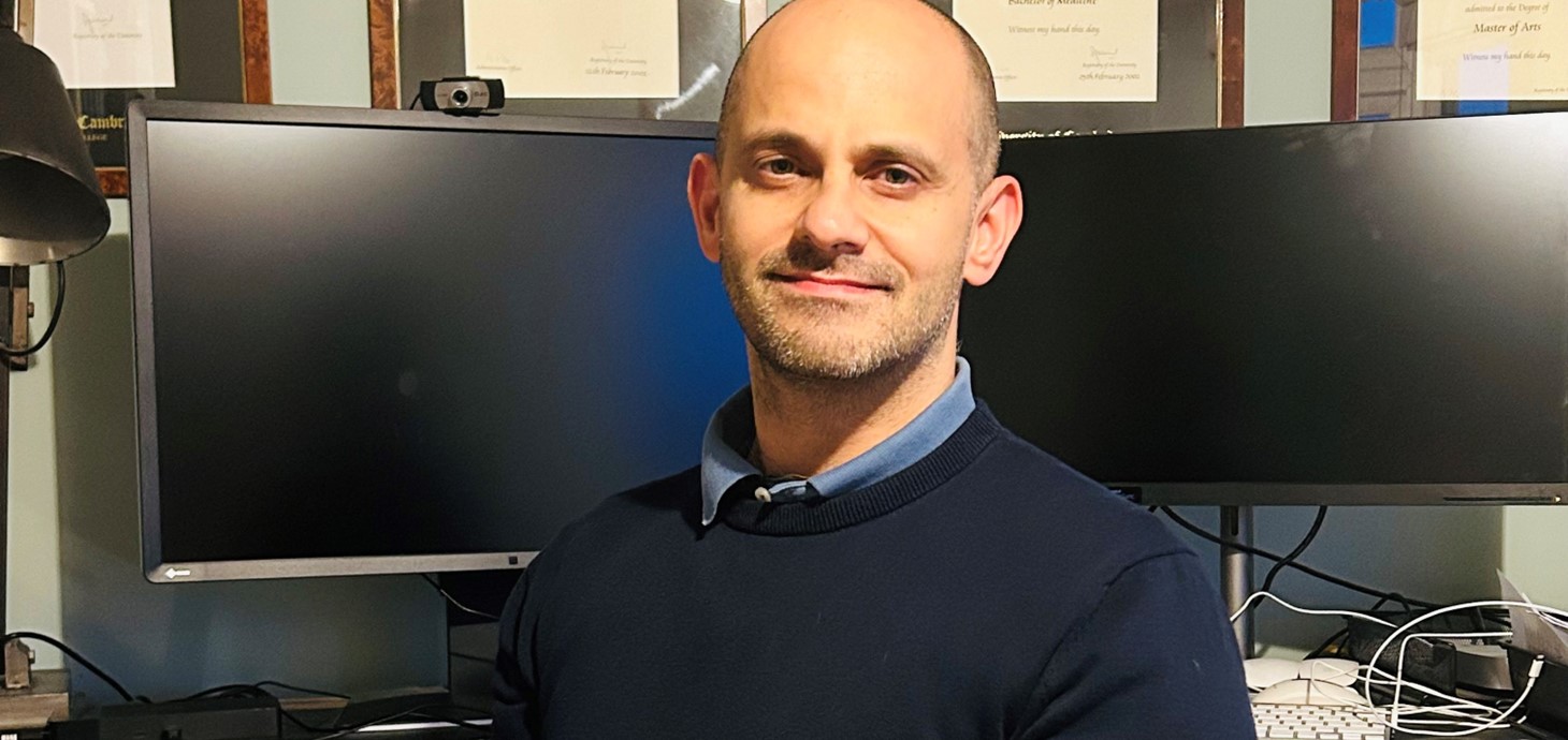 Dyn gwyn yn edrych ar gamera o flaen dau sgrin cyfrifiadur