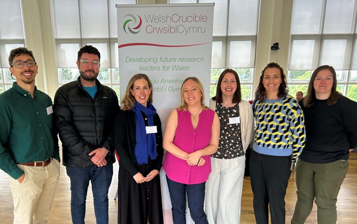 Llun grŵp o'r saith ymchwilydd o Brifysgol Abertawe'n sefyll gyda'i gilydd ac yn gwenu o flaen baner naid sy'n arddangos logo Crwsibl Cymru.