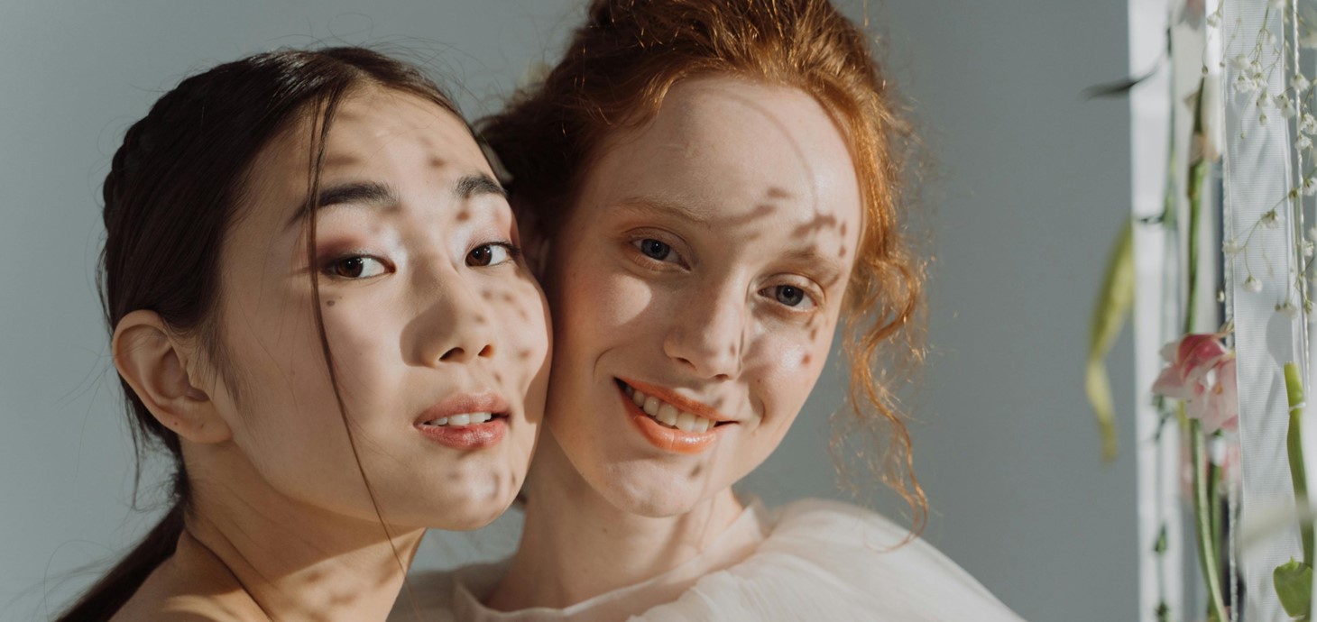 Dwy fenyw ifanc - un Asiaidd, un Cawcasaidd - yn sefyll boch wrth foch yn edrych ar y camera.