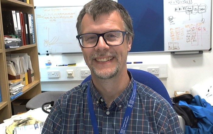 Dyn yn eistedd mewn swyddfa gyda bwrdd gwyn y tu ôl iddo