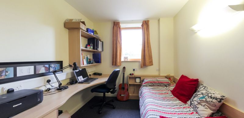 Ystafell wely i fyfyrwyr gyda gwely sengl, desg astudio, silffoedd a ffenestr.