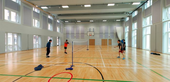 Pobl yn chwarae badminton mewn neuadd chwaraeon dan do llachar gyda chwrtiau pren.