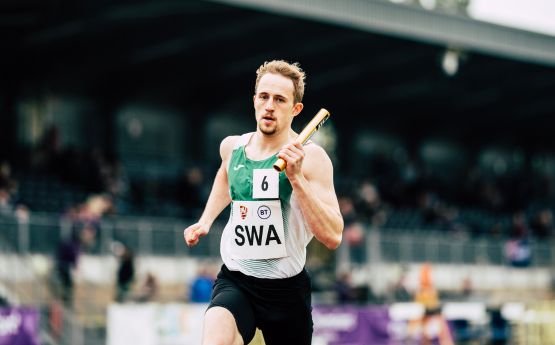 A Swansea University athelete running on the track in the middle of a relay race, holding baton.