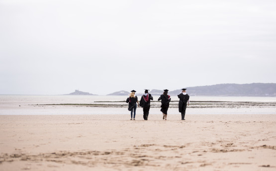 4 students in graduation robes seen walking in the distance on Swansea beach