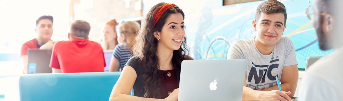 Two students working on a laptop whilst chatting to another student