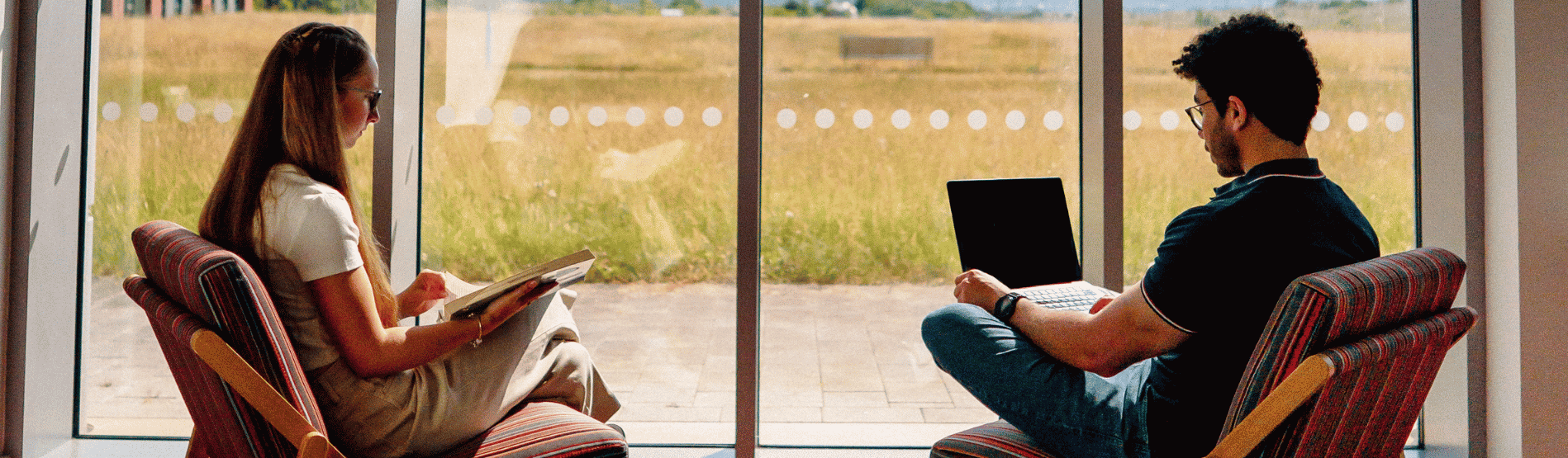 Two students sitting at the Bay Library, looking through the large glass windows with a book in hand.