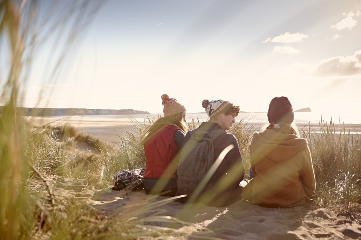 Two female students and one male student sitting in the sane dunes and looking out to the sea.