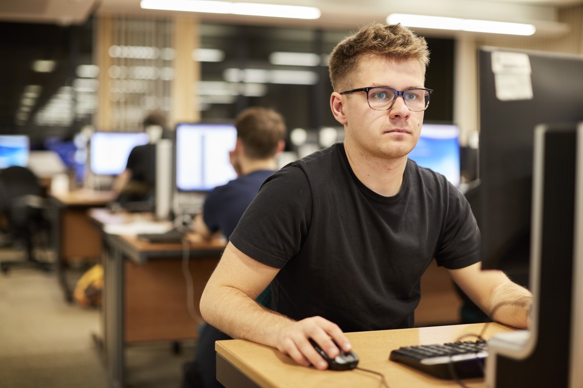 Male student wearing a black t-shirt and glasses looking sitting at a computer