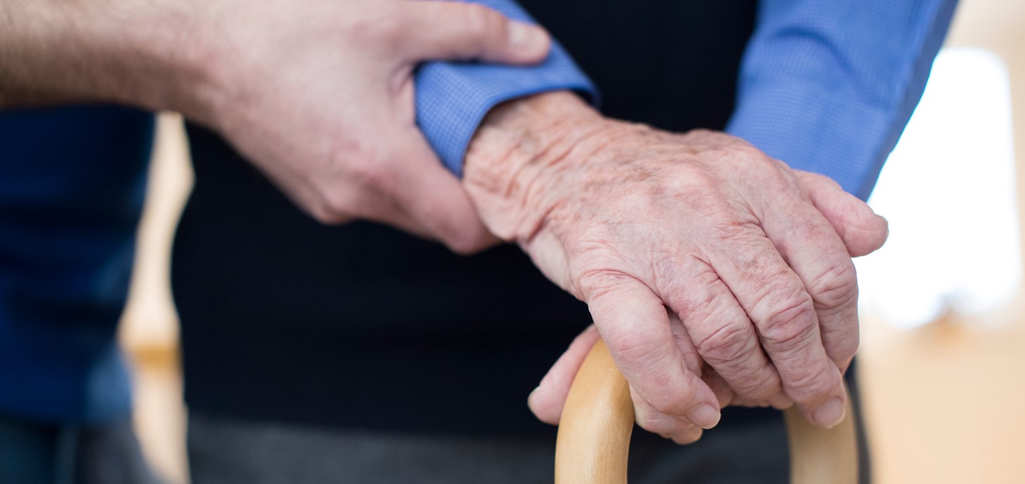 An elderly persons hands on a walking stick with another person's hand offering support
