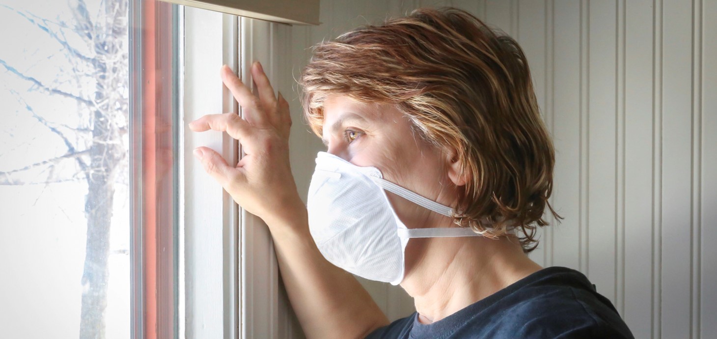 Woman wearing a mask looks sadly out the window of her home