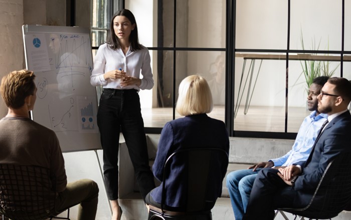 A woman standing in front of a group of seated people alongside a whiteboard