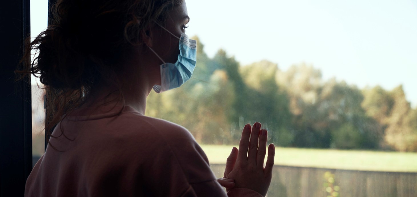 Back view of a woman with protective mask standing indoors looking out of a window.