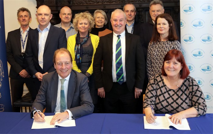 Man and woman sitting at table signing document with group of nine people standing behind them - all looking at the camera