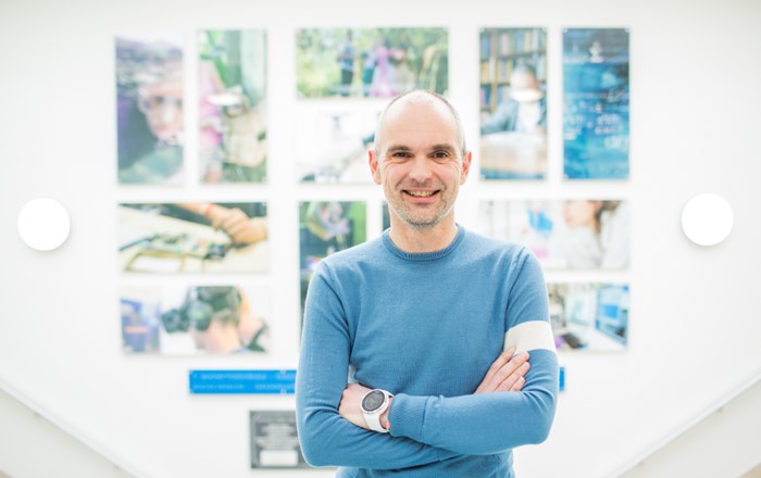 Professor Matt Jones is smiling and has his arms folded. He is wearing a blue sweater and standing in front of brightly coloured artwork.