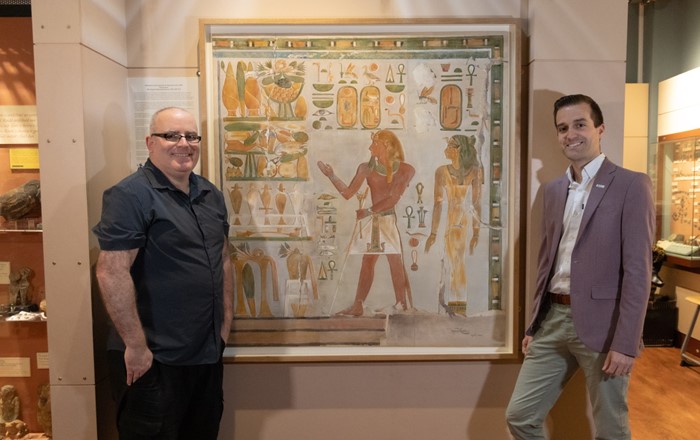Two men standing either side of a large Egyptian painting inside a gallery