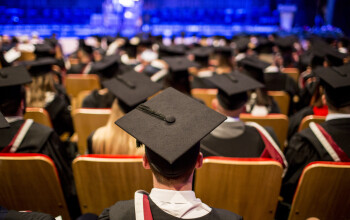 A graduation ceremony with graduates in caps and gowns sitting in rows, viewed from behind, with a stage in soft focus in the background.