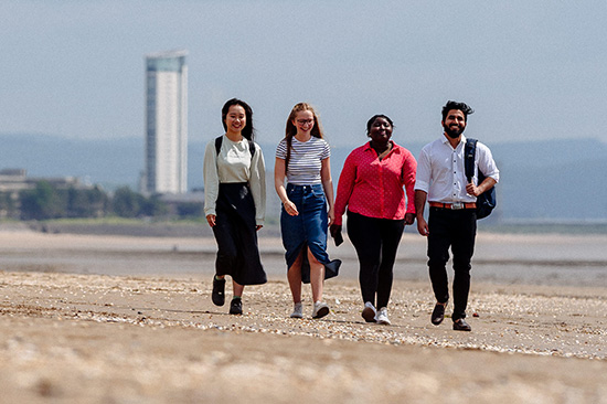Four students walking along Swansea beach, the iconic Meridian Tower in the background.