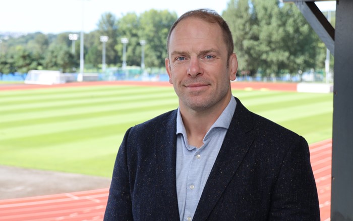 A headshot of Alun Wyn Jones wearing a suit. In the background is Swansea Bay Sports Park.