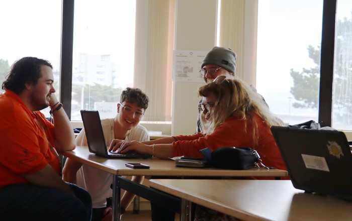 Four people are at desks in a classroom looking at laptop screens. 