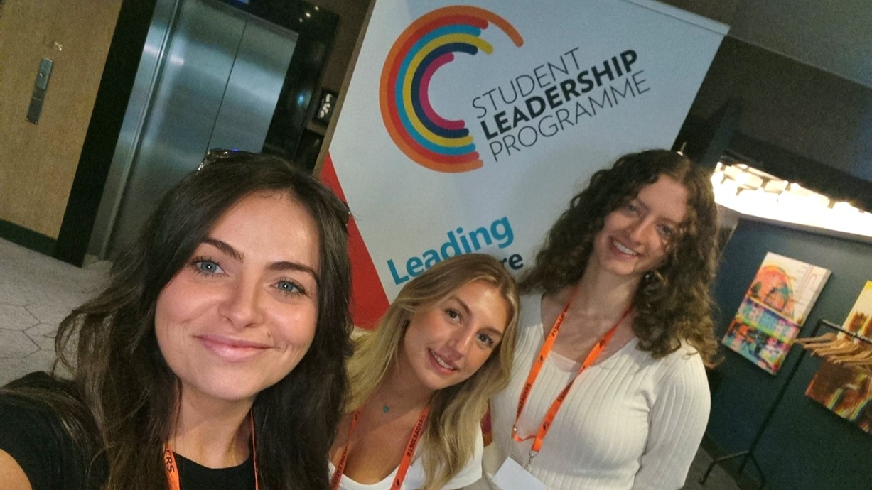 A selfie of Cerys, Lily, and Ellesse at a recent welcome event for the Student Leadership Programme. In the background is a pull-up banner with the programme’s logo on it.