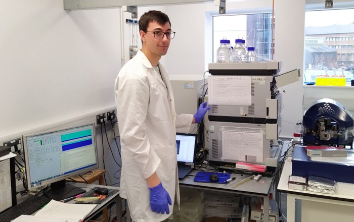 First-year SWBio student Tom wears a white lab coat and safety goggles as he works in the mass spectrometry lab.